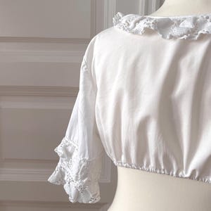 May include: A white, cropped blouse with a round, ruffled neckline and short sleeves. The sleeves feature lace trim at the cuffs. The blouse is displayed on a mannequin.