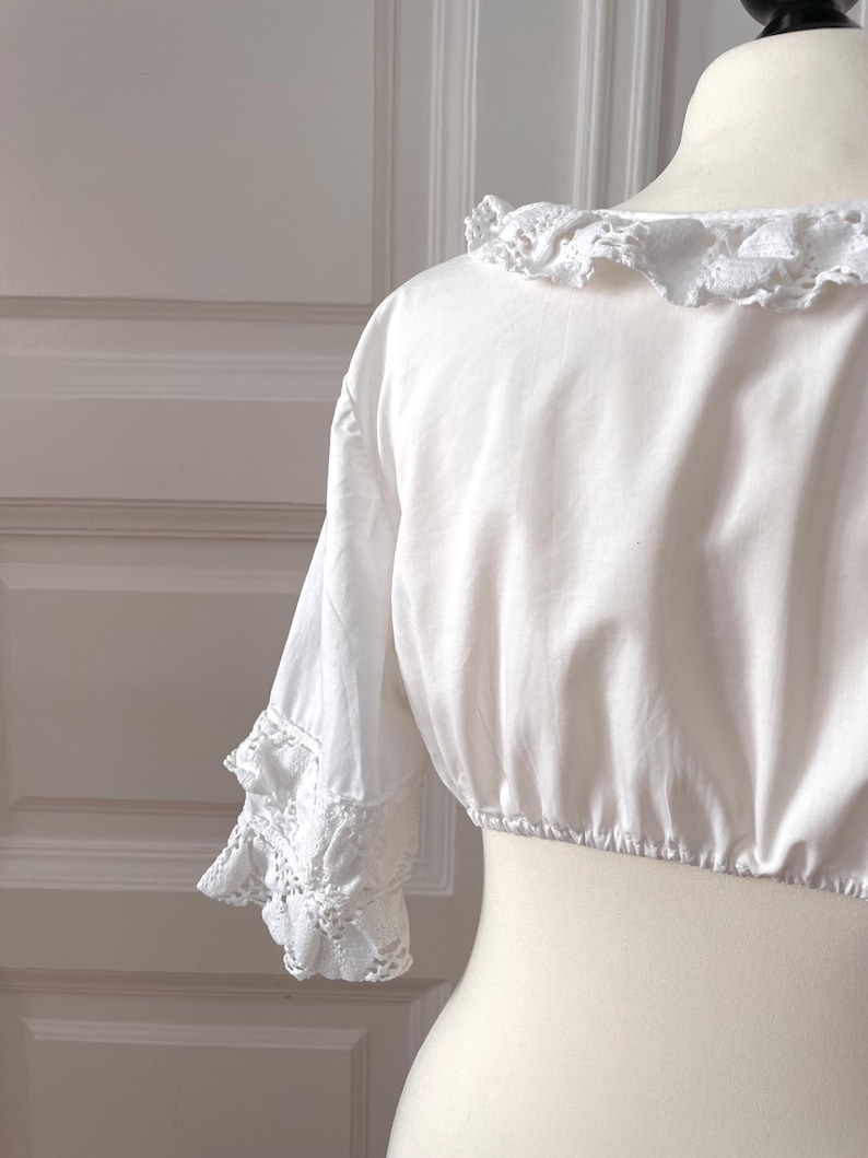 May include: A white, cropped blouse with a round, ruffled neckline and short sleeves. The sleeves feature lace trim at the cuffs. The blouse is displayed on a mannequin.