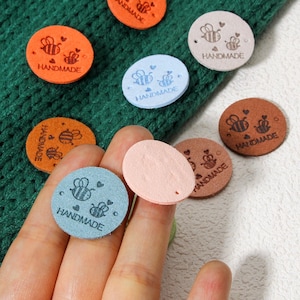 Op de afbeelding: Assortiment van ronde, handgemaakte viltlabels met een bijendesign. De labels zijn in verschillende kleuren, waaronder blauw, roze, bruin en lichtblauw. Elk label heeft de tekst "HANDMADE" erop gedrukt.