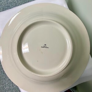 May include: Off-white ceramic plate with a wide rim. The underside features a circular base and a central area with the text "Made in PORTUGAL". The plate has a simple, elegant design, suitable for various dining settings.