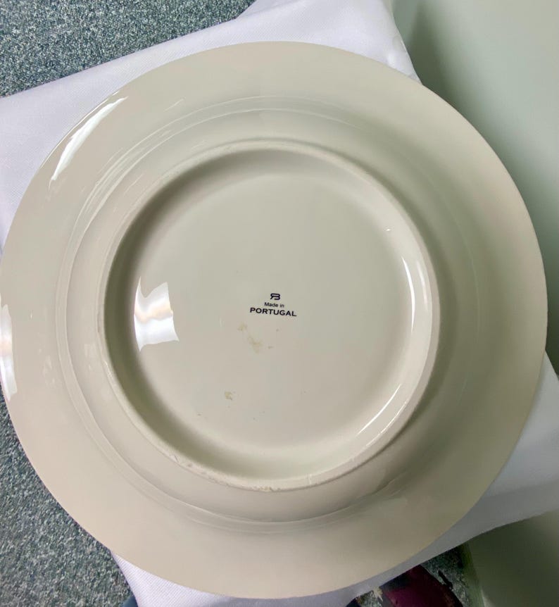 May include: Off-white ceramic plate with a wide rim. The underside features a circular base and a central area with the text "Made in PORTUGAL". The plate has a simple, elegant design, suitable for various dining settings.