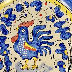 May include: A hand-painted ceramic plate featuring a blue rooster with a red comb and yellow feet. The plate has a white background with blue floral and leaf designs, a yellow border, and a blue rim.