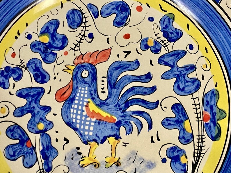 May include: A hand-painted ceramic plate featuring a blue rooster with a red comb and yellow feet. The plate has a white background with blue floral and leaf designs, a yellow border, and a blue rim.