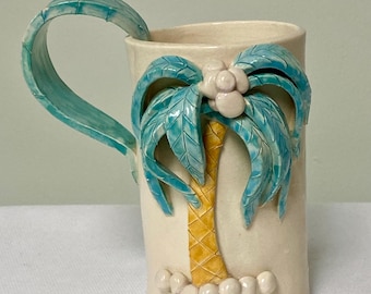 May include: A white ceramic mug with a 3D palm tree design. The palm tree has blue leaves and a yellow trunk. The mug has a blue handle.