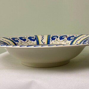 May include: A white ceramic serving bowl with a wide rim. The rim is decorated with blue floral patterns, yellow and red accents, and a blue dotted border. The bowl has a shallow, rounded shape.