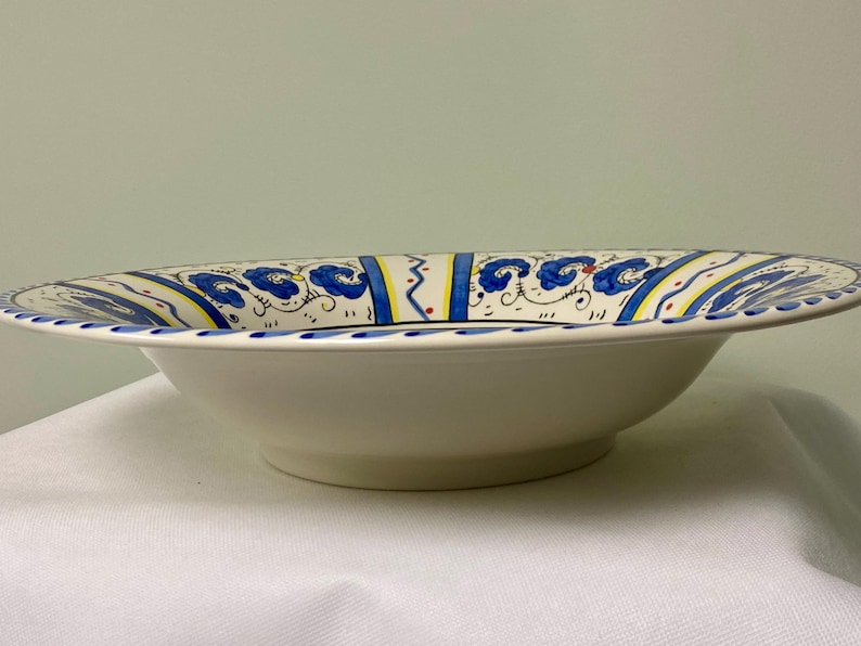 May include: A white ceramic serving bowl with a wide rim. The rim is decorated with blue floral patterns, yellow and red accents, and a blue dotted border. The bowl has a shallow, rounded shape.