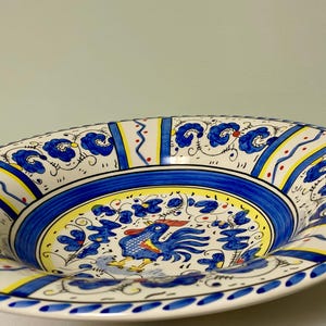 May include: A decorative ceramic serving bowl with a vibrant hand-painted design. The bowl features a blue and yellow color scheme with a central image of a rooster. The rim is adorned with floral and geometric patterns.