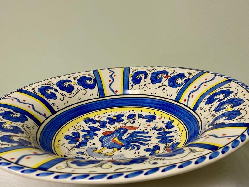 May include: A decorative ceramic serving bowl with a vibrant hand-painted design. The bowl features a blue and yellow color scheme with a central image of a rooster. The rim is adorned with floral and geometric patterns.