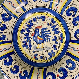 May include: A decorative ceramic plate with a rooster design. The plate features a blue, yellow, and white color scheme with floral and geometric patterns. The rooster is blue with red accents, and the plate has a scalloped edge.