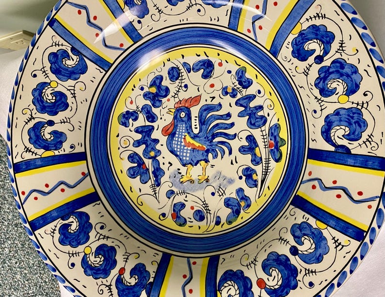 May include: A decorative ceramic plate with a rooster design. The plate features a blue, yellow, and white color scheme with floral and geometric patterns. The rooster is blue with red accents, and the plate has a scalloped edge.
