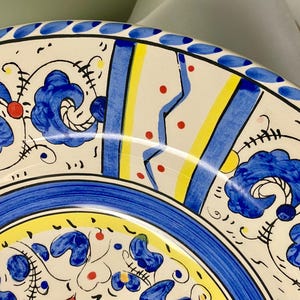 May include: Close-up of a decorative ceramic plate with a vibrant, hand-painted design. The plate features a white base with blue floral motifs, yellow and blue stripes, and red accents. A blue border with small white dots frames the edge.