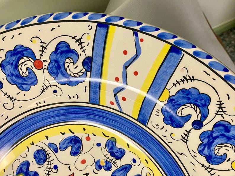 May include: Close-up of a decorative ceramic plate with a vibrant, hand-painted design. The plate features a white base with blue floral motifs, yellow and blue stripes, and red accents. A blue border with small white dots frames the edge.
