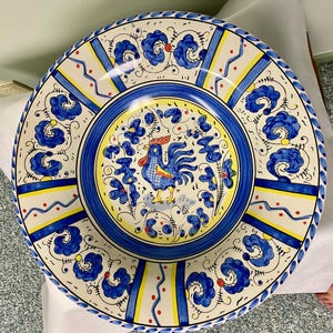 May include: A large, round ceramic plate with a hand-painted design. The plate features a blue and yellow color scheme with a central image of a rooster. The rim has a blue floral pattern, and the plate is approximately 12 inches in diameter.