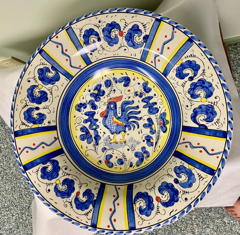 May include: A large, round ceramic plate with a hand-painted design. The plate features a blue and yellow color scheme with a central image of a rooster. The rim has a blue floral pattern, and the plate is approximately 12 inches in diameter.