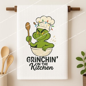 May include: White kitchen towel with a cartoon Grinch character wearing a chef's hat and holding a wooden spoon. The Grinch is in a bowl with the text "Grinchin' in the Kitchen".