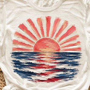 May include: White t-shirt with a sunset design. The artwork shows a sun with red and orange rays above blue and red water. The design has a textured, painted appearance, creating a unique look. The shirt is made of a soft, comfortable material.