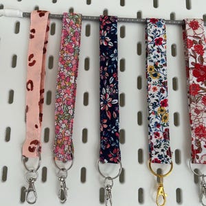 Puede incluir: Seis cordones de tela con diferentes diseños florales y de animales. Los cordones presentan tonos rosas, azules y rojos, cada uno con un cierre metálico. Se exhiben en un panel perforado blanco.
