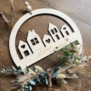 May include: A wooden circle with a cutout design of houses and the text "Zuhause ist es am schönsten" in a cursive font. The circle is decorated with dried eucalyptus and pampas grass.