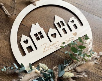 Puede incluir: Un círculo de madera con un diseño recortado de casas y el texto "Zuhause ist es am schönsten" en una fuente cursiva. El círculo está decorado con eucalipto seco y hierba de la pampa.