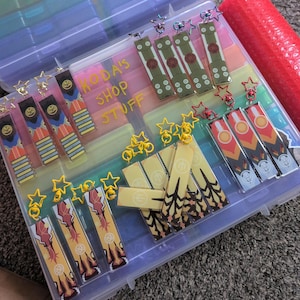 May include: A clear plastic storage container filled with various keychains and charms. The keychains feature colorful designs with star-shaped and circular accents. The text "MODA'S SHOP STUFF" is visible on the dividers.