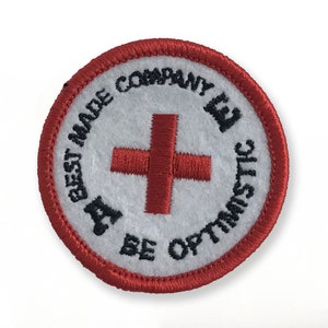 May include: Red embroidered patch with a white background and the text "A Best Made Company Be Optimistic" in black. A red cross is in the center of the patch.