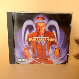 Op de afbeelding: Een cd-hoesje met het album "Spare Ass Annie and Other Tales" van William S. Burroughs. De hoes toont een man in pak en hoed tegen een paars-witte achtergrond. Een waarschuwing is zichtbaar.