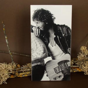 Puede incluir: Impresión en blanco y negro de un músico con cabello rizado, que lleva una chaqueta de cuero y sostiene una guitarra. La imagen es un retrato, con el músico apoyado en otra persona. La guitarra es de color claro con un golpeador oscuro.