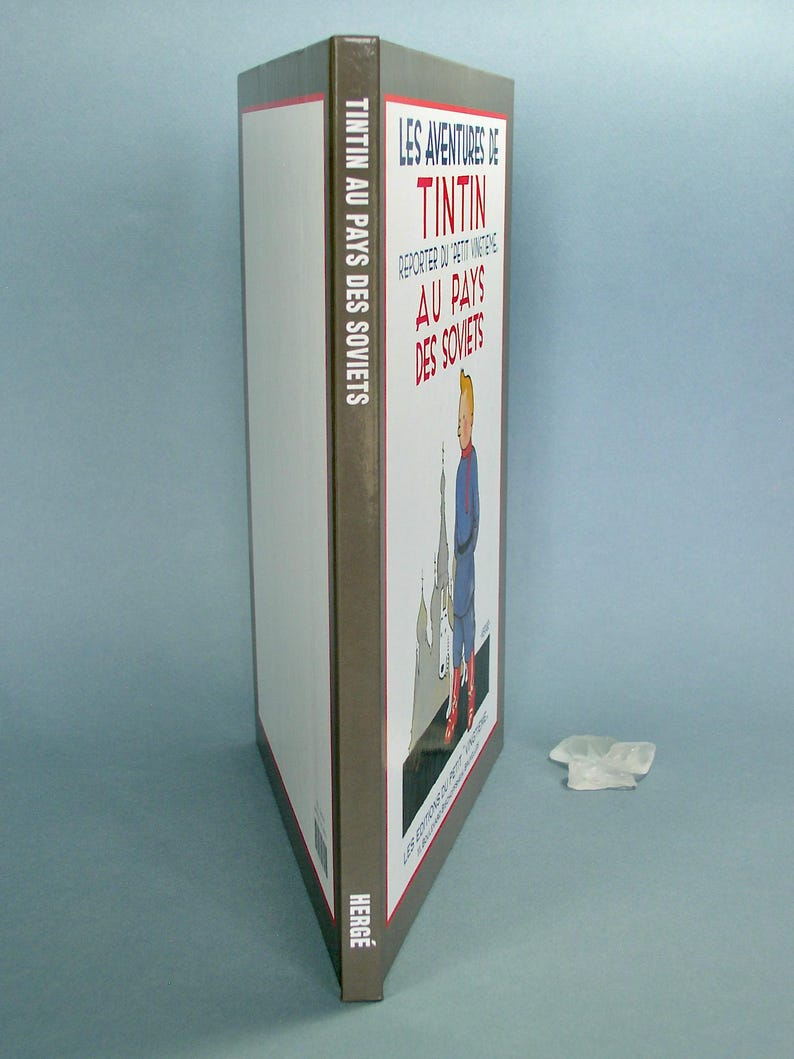 Może przedstawiać: Szara książka w twardej oprawie z tytułem "Les Aventures de Tintin Reporter au Petit Vingti&egrave;me Au Pays des Soviets" i autorem "Herg&eacute;" na grzbiecie. Okładka przedstawia rysunkową ilustrację Tintina stojącego przed budynkiem z czerwoną gwiazdą na szczycie.