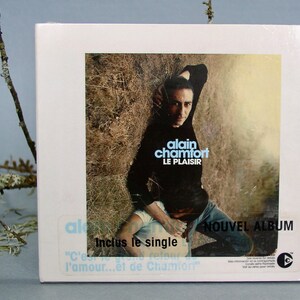 Peut inclure: Une pochette de CD blanche avec une photo d'un homme portant une chemise noire et un jean bleu. Le texte sur la pochette indique "alain chamfort LE PLAISIR" et "NOUVEL ALBUM".