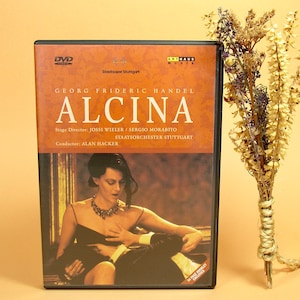 Peut inclure: Pochette de DVD pour l'opéra "Alcina" de Georg Friedrich Händel. La pochette présente une femme en robe noire avec un collier, et le titre de l'opéra est écrit en gros caractères gras.
