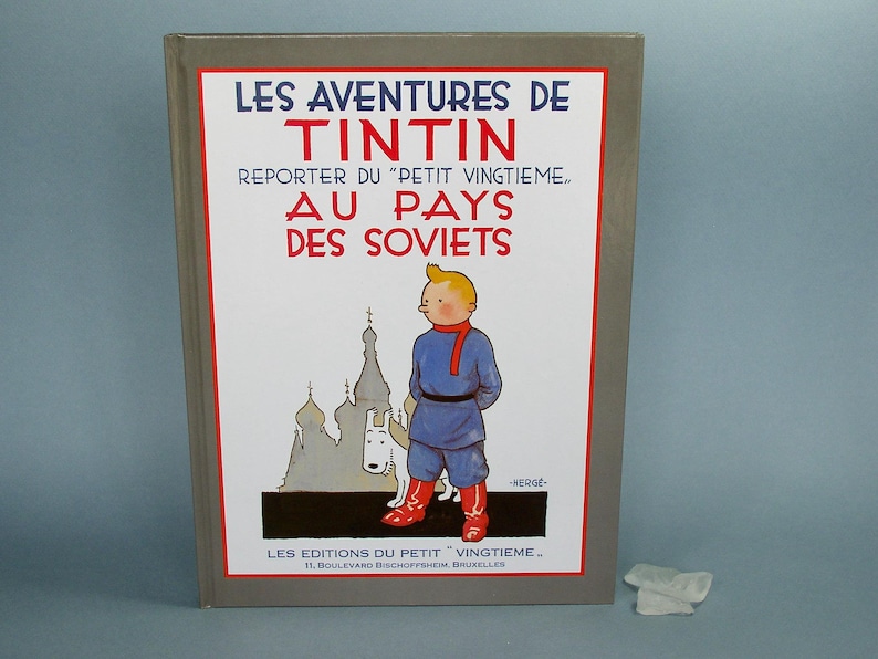 Może przedstawiać: Antyczna książka w twardej oprawie zatytułowana "Les Aventures de Tintin Reporter du "Petit Vingtieme." Au Pays des Soviets" z ilustracją Tintina i Milusia przed budynkiem. Okładka książki jest szara z czerwonym i białym tekstem.