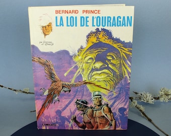 May include: A vintage book titled "LA LOI DE L'OURAGAN" by Bernard Prince. The cover features a colorful illustration of a parrot, a large face, and figures with spears. The book has a white spine and the publisher's logo.