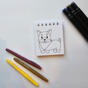 May include: A white notepad with a black and white line drawing of a dog on it. There are four markers next to the notepad: one brown, one yellow, one light yellow, and one dark red.