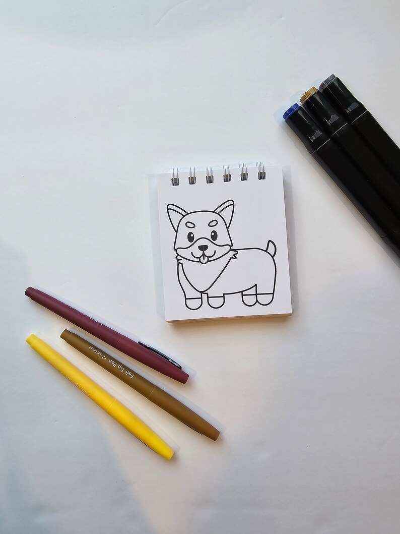 May include: A white notepad with a black and white line drawing of a dog on it. There are four markers next to the notepad: one brown, one yellow, one light yellow, and one dark red.