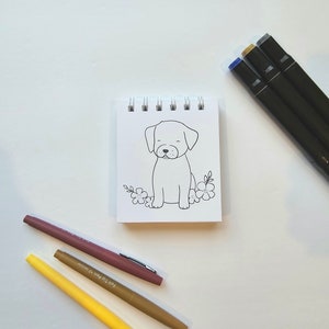 May include: A small, white notepad with a black and white line drawing of a dog sitting with flowers. The notepad is surrounded by three markers: one brown, one yellow, and one pink.