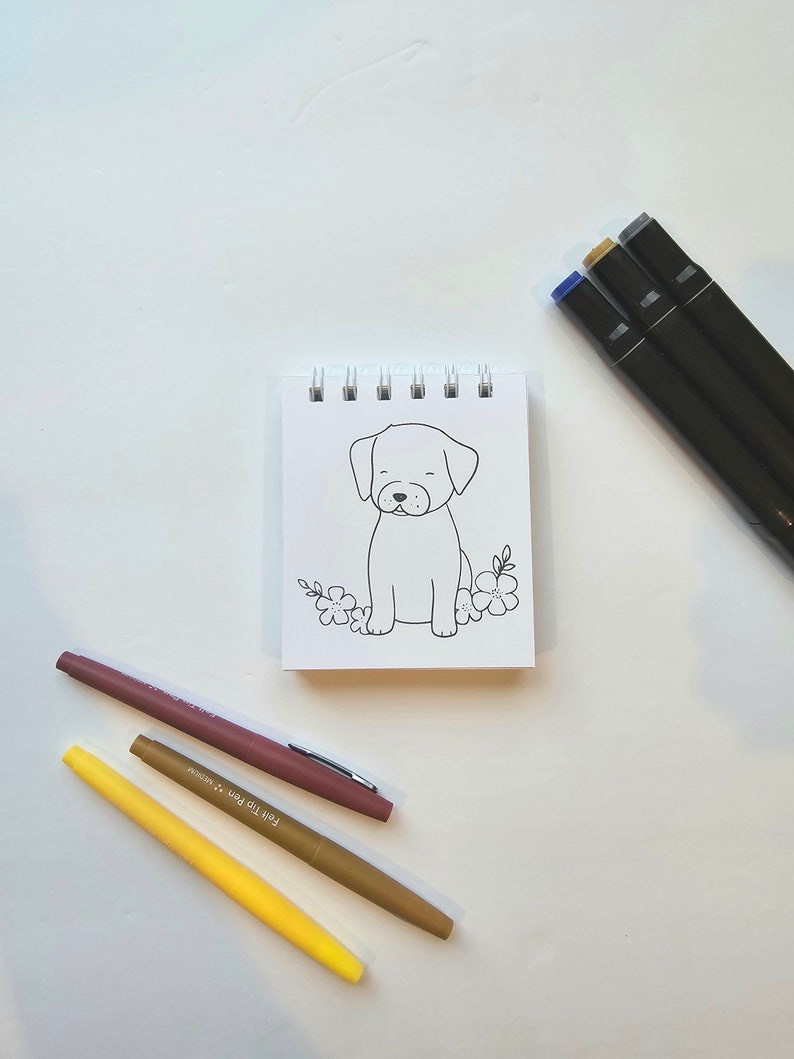 May include: A small, white notepad with a black and white line drawing of a dog sitting with flowers. The notepad is surrounded by three markers: one brown, one yellow, and one pink.
