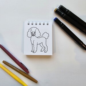 May include: A white, line-drawn illustration of a poodle on a small, white notepad. The notepad is surrounded by three markers: one brown, one yellow, and one black.