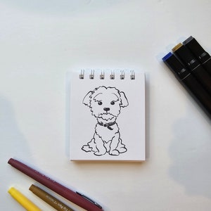 May include: A black and white line drawing of a dog sitting on a white notepad. The dog has a collar and is looking at the viewer. The notepad is on a white surface. There are three colored markers on the surface.