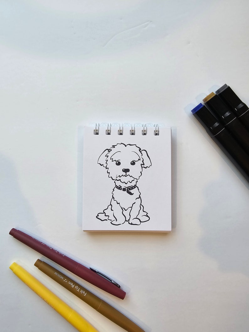 May include: A black and white line drawing of a dog sitting on a white notepad. The dog has a collar and is looking at the viewer. The notepad is on a white surface. There are three colored markers on the surface.