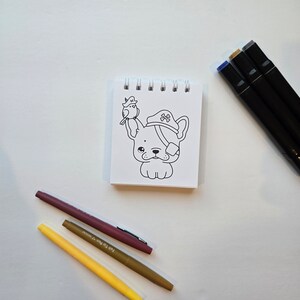 May include: A white notepad with a black and white line drawing of a dog wearing a pirate hat and a bird on its head. The notepad is surrounded by three markers: a yellow marker, a brown marker, and a blue marker.