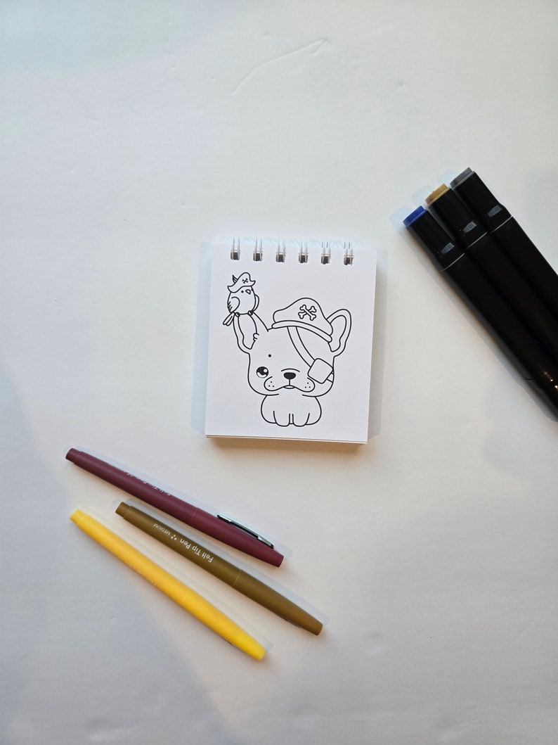May include: A white notepad with a black and white line drawing of a dog wearing a pirate hat and a bird on its head. The notepad is surrounded by three markers: a yellow marker, a brown marker, and a blue marker.