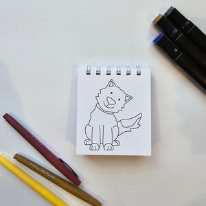 May include: A black and white line drawing of a dog sitting on a white sheet of paper in a spiral bound notebook. The dog is smiling and has a tail. There are three colored markers on a white surface below the notebook.