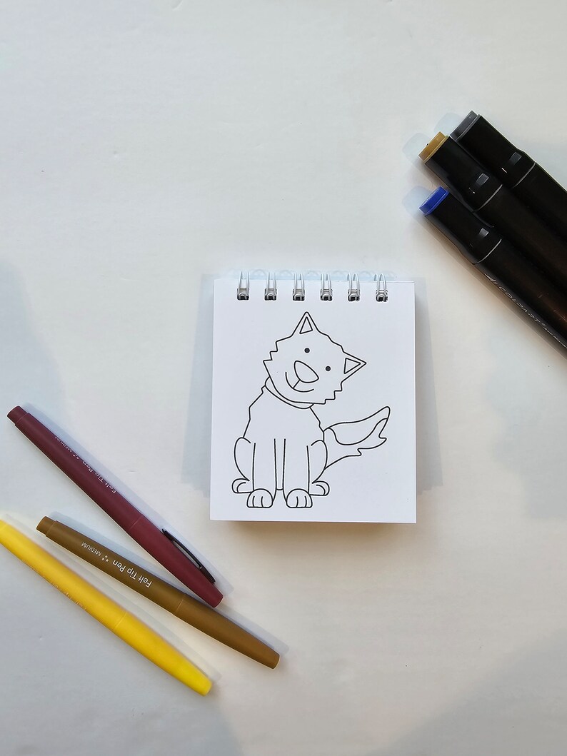 May include: A black and white line drawing of a dog sitting on a white sheet of paper in a spiral bound notebook. The dog is smiling and has a tail. There are three colored markers on a white surface below the notebook.