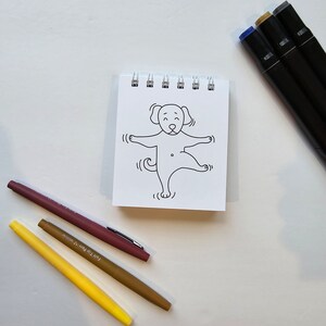 May include: A small, white notepad with a black and white line drawing of a dog on the front. The dog is standing on its hind legs with its front paws raised. There are three markers on the table: a brown marker, a yellow marker, and a black marker.
