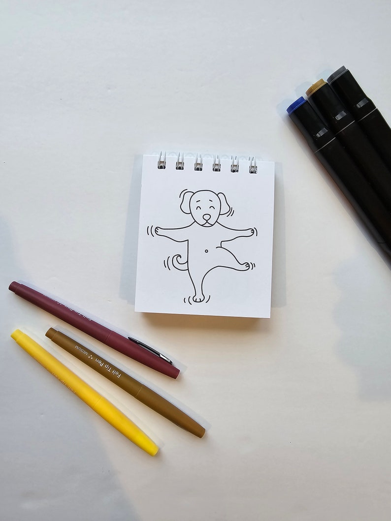 May include: A small, white notepad with a black and white line drawing of a dog on the front. The dog is standing on its hind legs with its front paws raised. There are three markers on the table: a brown marker, a yellow marker, and a black marker.