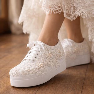 May include: White platform sneakers featuring intricate lace overlays and small pearl embellishments. The shoes have white laces, a thick rubber sole, and a floral-patterned lace design covering the upper portion of the footwear.