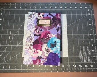 May include: A floral journal with a purple background and a variety of flowers in shades of pink, purple, and blue. The journal has a lace border on the left side and a metal label holder.
