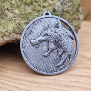May include: A silver-coloured metal pendant featuring a detailed wolf head design. The wolf's mouth is open, and its teeth are visible. The pendant is circular and has a textured surface.