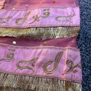 May include: Close-up of a pink fabric with gold accents. The fabric has a sheer, dotted overlay and is trimmed with gold fringe. Gold embroidered text is visible on the pink bands. The overall aesthetic suggests a decorative textile.