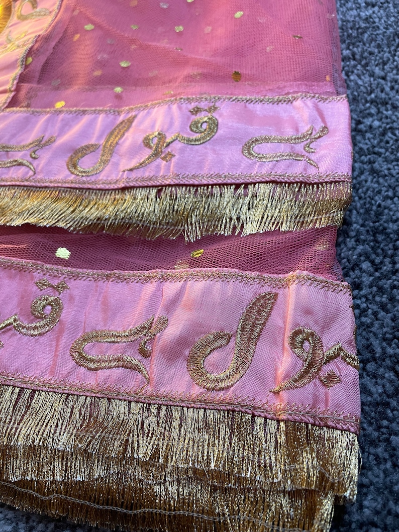 May include: Close-up of a pink fabric with gold accents. The fabric has a sheer, dotted overlay and is trimmed with gold fringe. Gold embroidered text is visible on the pink bands. The overall aesthetic suggests a decorative textile.